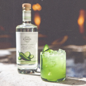 A green Cool as a Cucumber cocktail sitting in front of a bottle of 21Seeds Cucumber Jalapeño