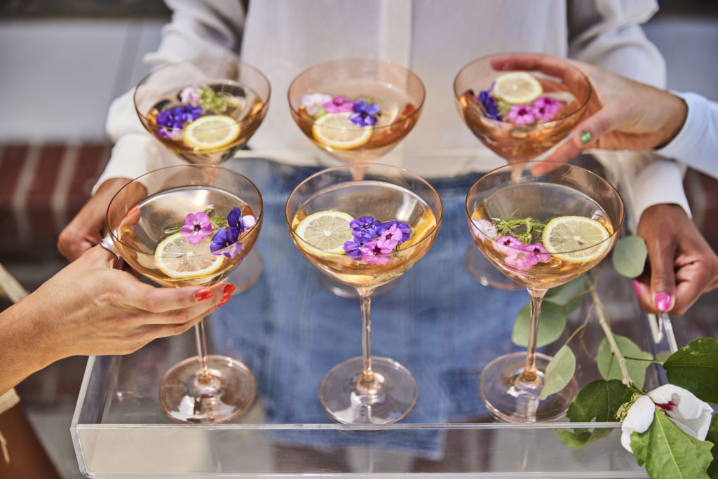 Six cocktails on a serving tray. There are two hands holding the glasses.