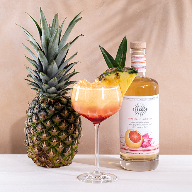 21Seeds Sayulita Sunrise cocktail garnished with a pineapple slice, sitting in front of a pineapple and a bottle of 21Seeds Grapefruit Hibiscus Infused Tequila