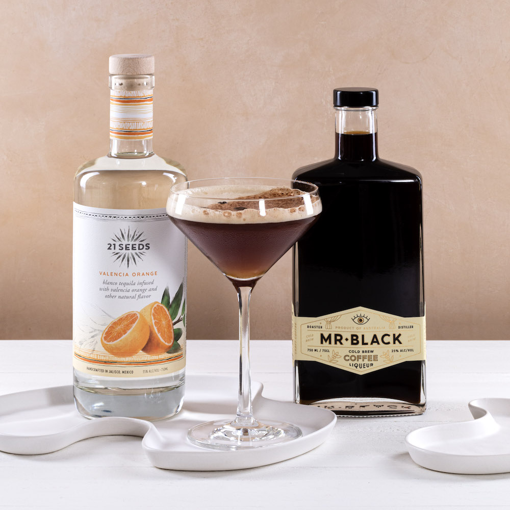 A 21Seeds Espresso Martini cocktail sitting on a table in front of a bottle of 21Seeds Valencia Orange Infused Tequila and a bottle of Mr Black Coffee Liqueur