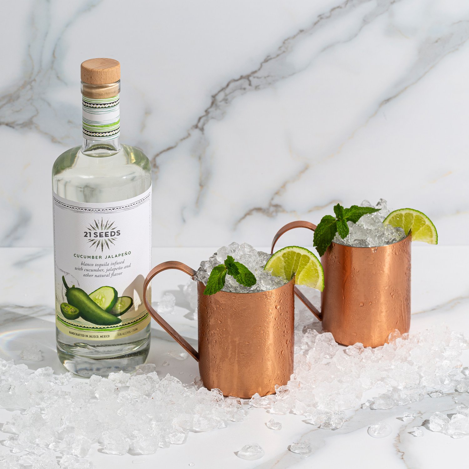 Two Jalisco mule cocktails in copper mugs garnished with a lime wheel and a mint sprig on a ice covered table. A bottle of 21Seeds Cucumber Jalapeño sits to the left of the cocktails.