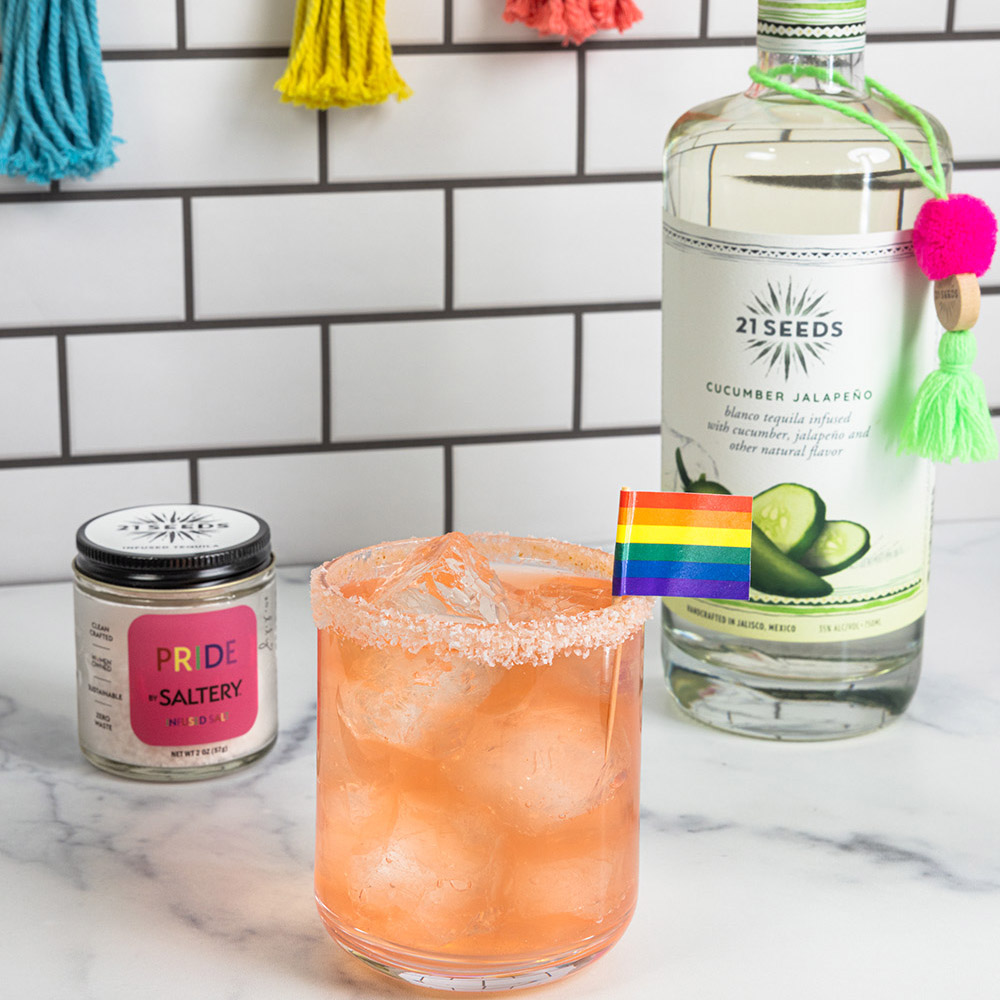 The proud & spicy cocktail on a table, garnished with a mini pride flag. A bottle of 21Seeds Cucumber Jalapeño Infused Tequila is in the background.