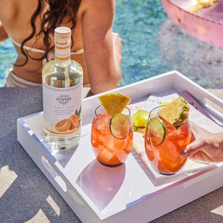 Two 21Seeds Tequila on the Beach cocktails on a serving tray with a bottle of 21Seeds Valencia Orange Infused tequila. The serving tray is poolside with a person standing in the pool in the background. 